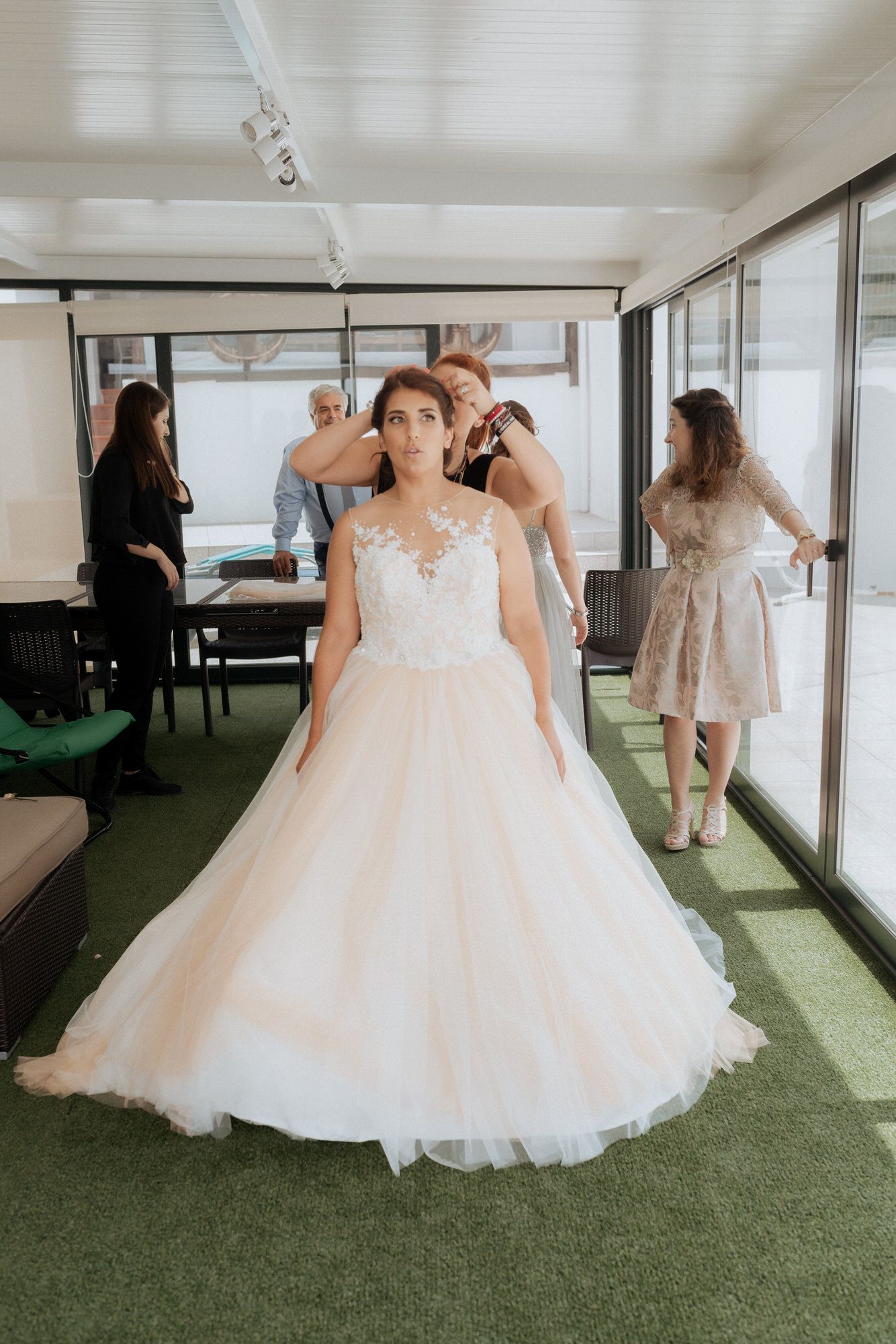 A bride getting ready in a stunning tulle ball gown with illusion neckline while others assist in a bright sunlit room.
