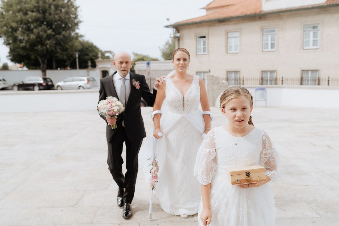Wedding attendees walk together on a sun-lit stone courtyard, with the bride wearing a white gown and others in formal attire.