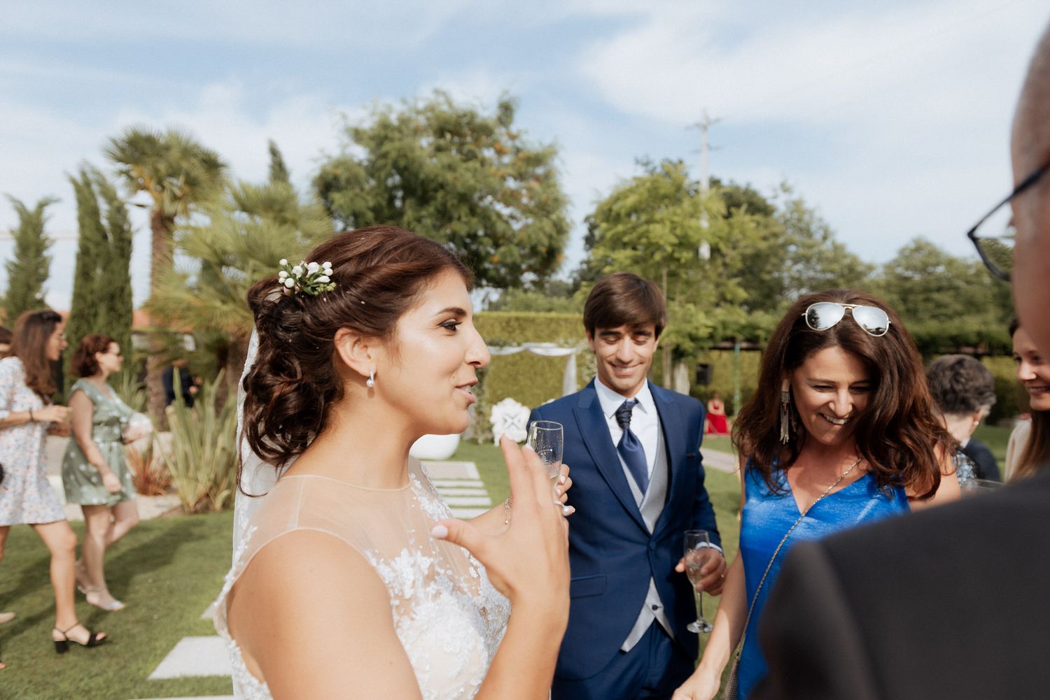 A group of wedding guests enjoying drinks and conversation outdoors on a sunny day in a garden setting.