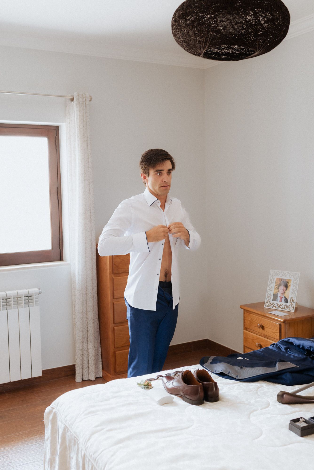 Person getting dressed in a bedroom with clothes laid out on white bed and wooden furniture.