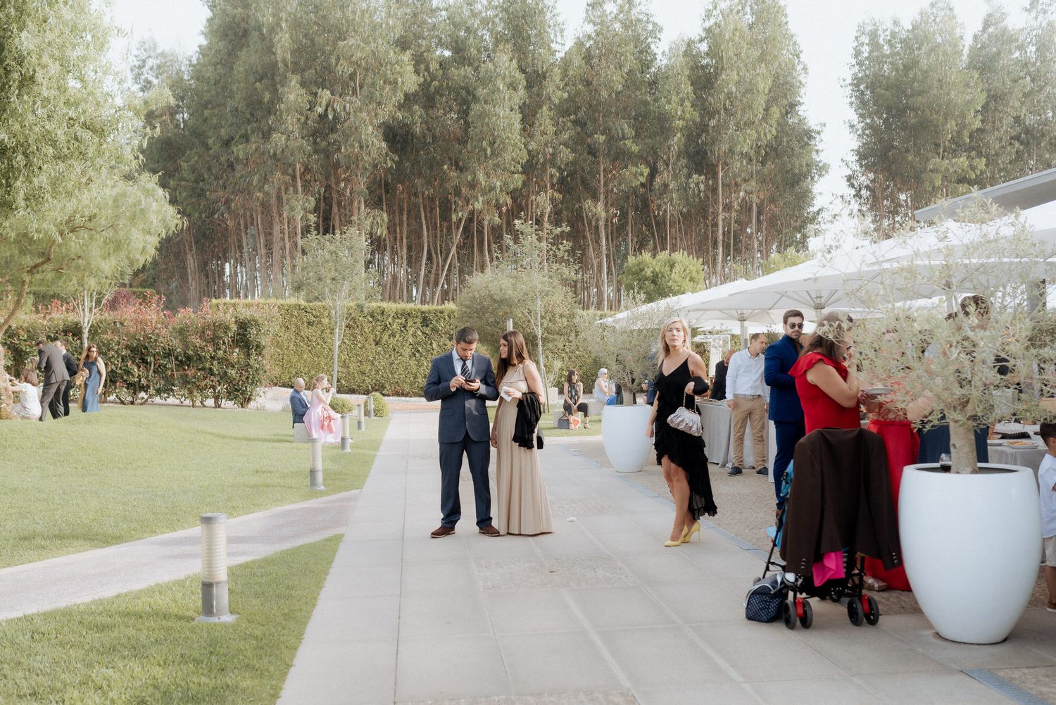 Wedding guests gather on a paved walkway surrounded by green trees and white decorative elements at an outdoor venue.