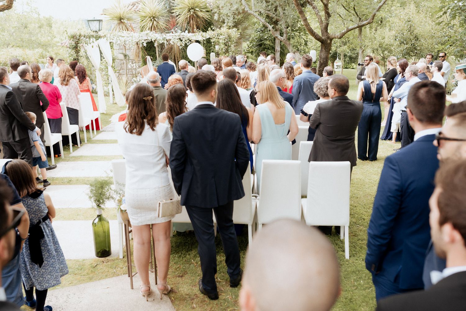 Wedding guests in formal attire stand outdoors under trees watching a ceremony at a garden venue.