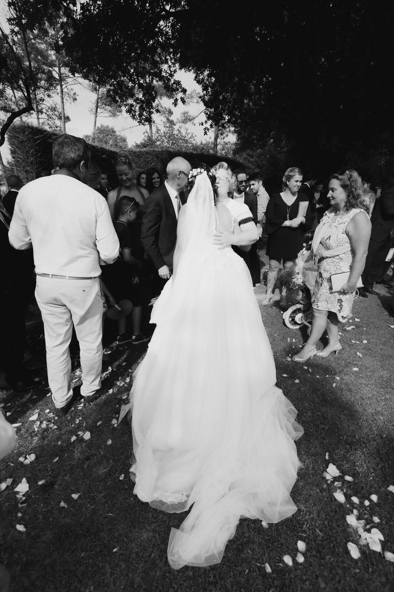 Black and white photo of a bride in a flowing white gown surrounded by guests at an outdoor wedding ceremony.