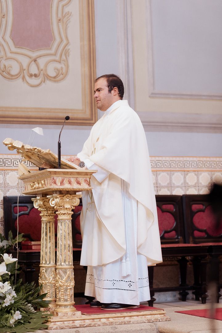 A priest in white vestments delivers a sermon from an ornate gold-detailed pulpit in an elegant church setting.