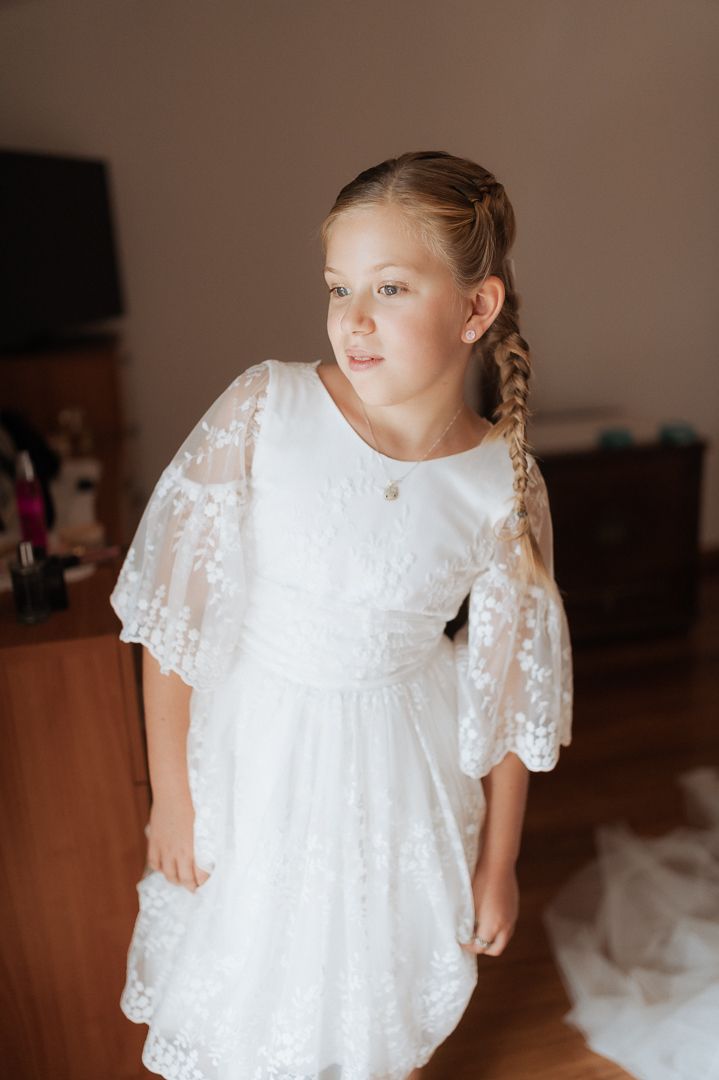 A child wearing a delicate white lace dress with sheer sleeves stands in a room during a formal occasion.