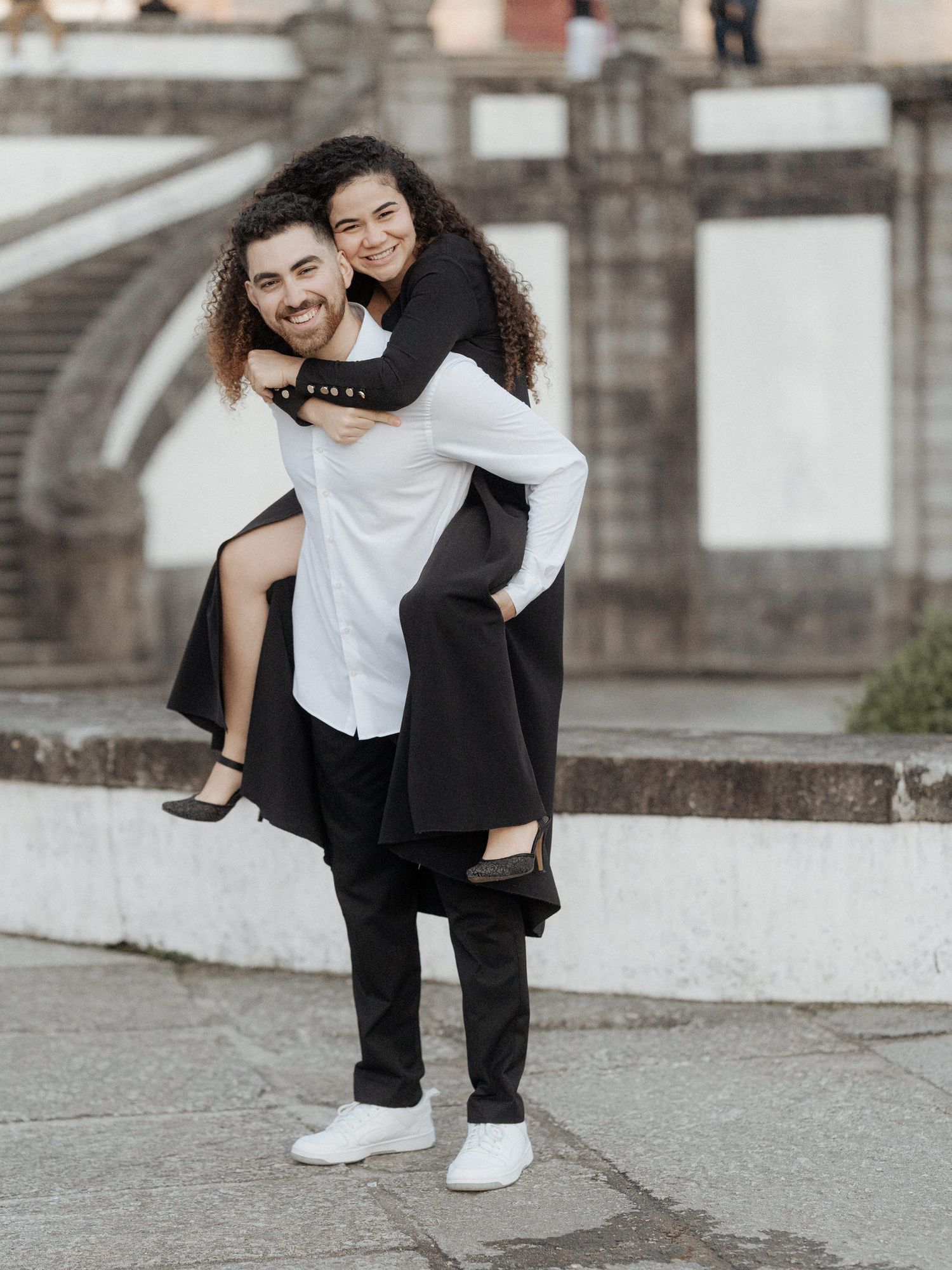 Happy couple in matching formal wear share a playful piggyback ride outdoors against architectural backdrop.