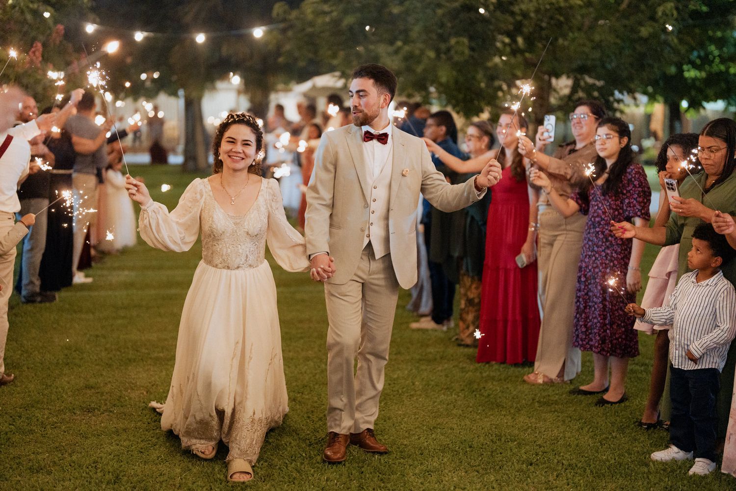 Couple walks between rows of guests holding sparklers during outdoor evening wedding reception with string lights overhead.