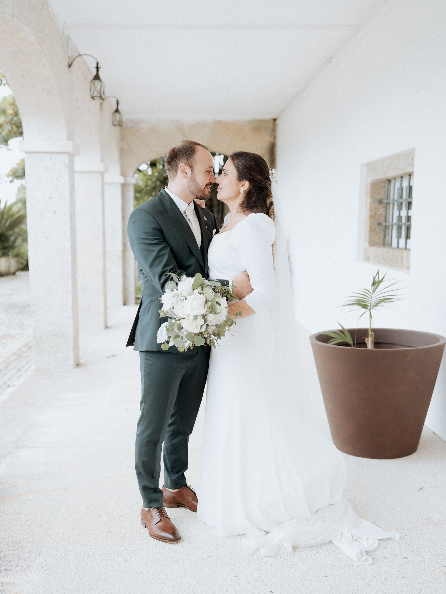 Newlywed couple in elegant attire share intimate moment on white veranda with potted plant.