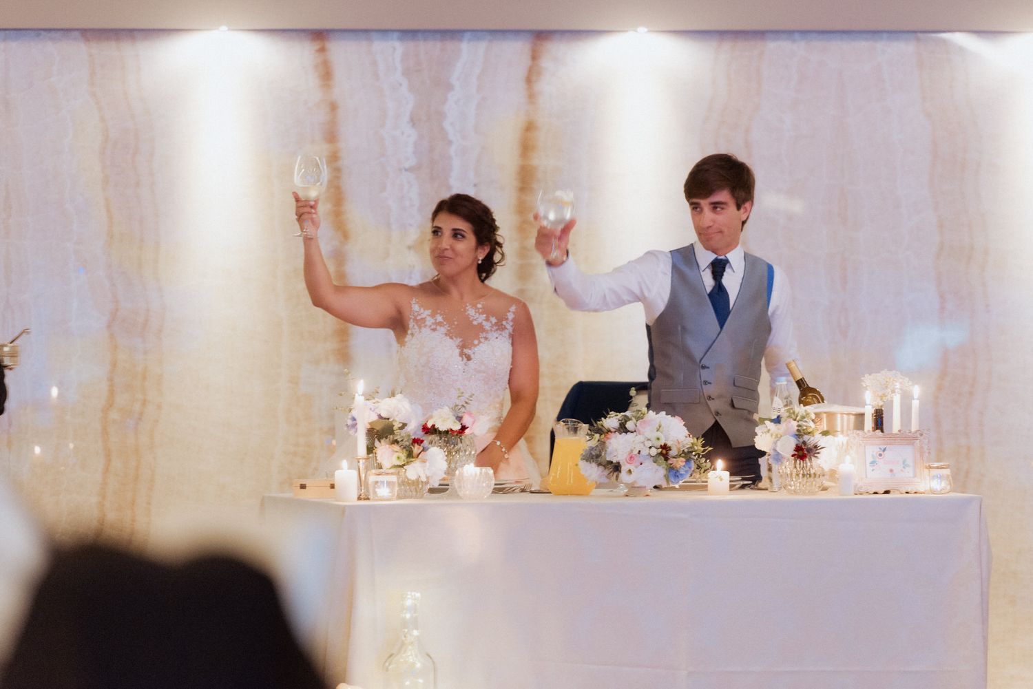 Wedding party raises their glasses in celebration during wedding reception toasts at elegant white-decorated venue.
