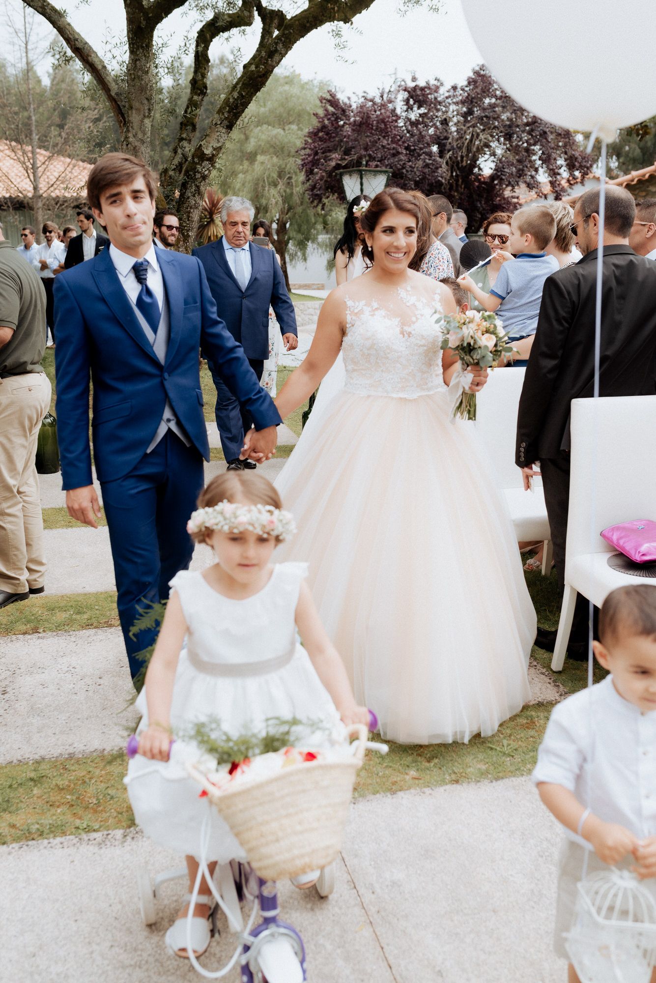 A wedding party in blue suits and white dresses gathers outdoors near trees on a sunny day.