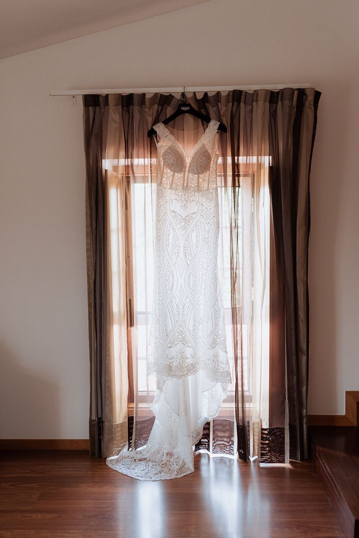 A white lace wedding dress hangs elegantly against sheer brown curtains in front of a sunlit window.