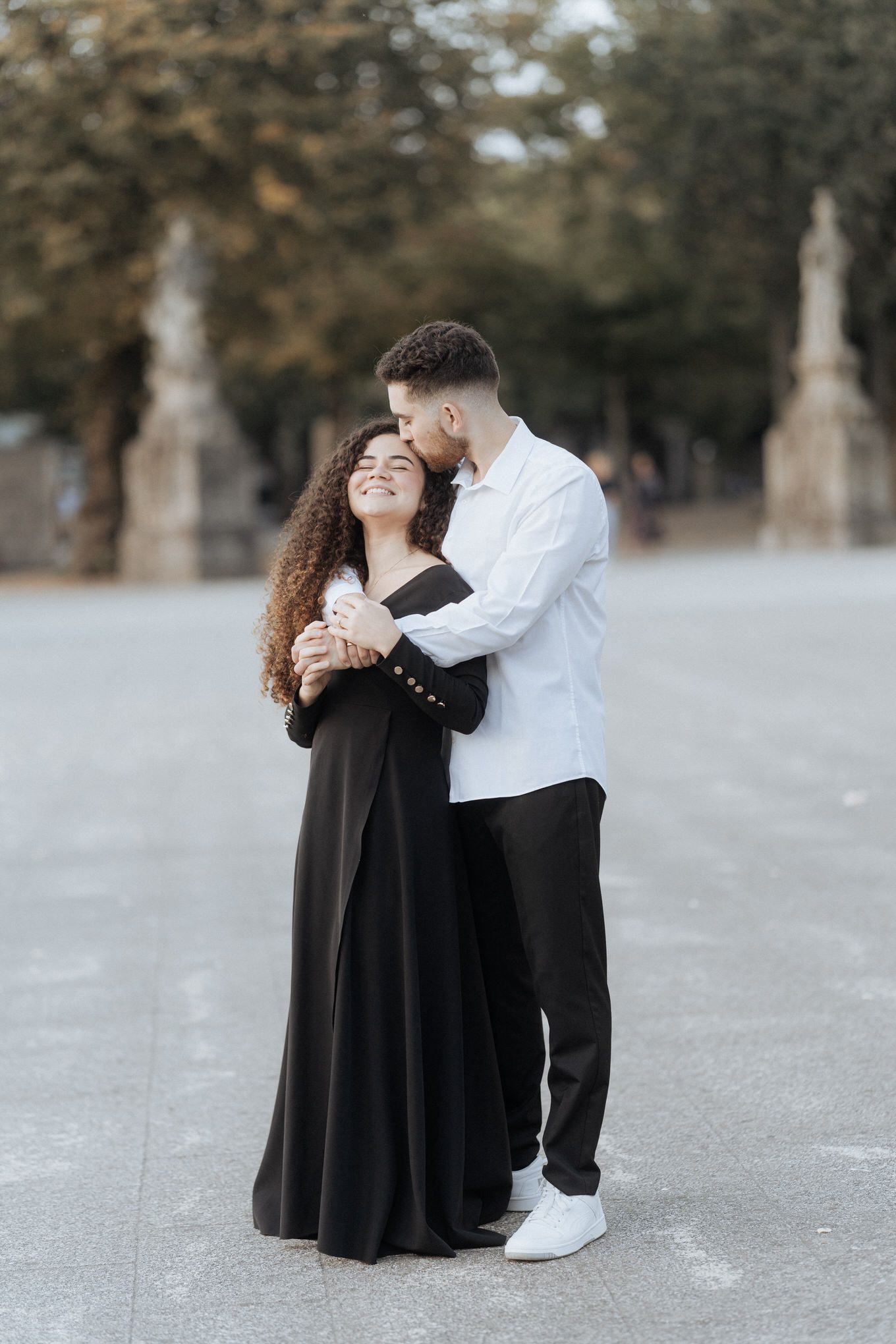 Um casal romântico se abraça ao ar livre, com a pessoa de preto usando um vestido esvoaçante e a outra com uma camisa branca.