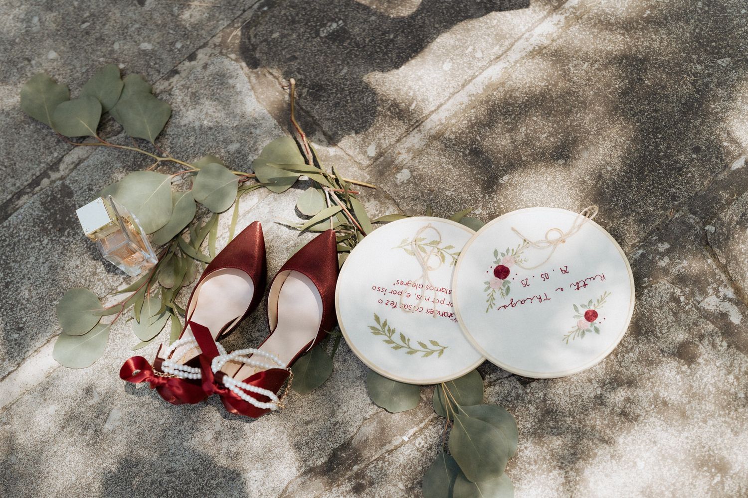 Red wedding shoes and RSVP cards styled with eucalyptus leaves on a textured stone surface in natural lighting.