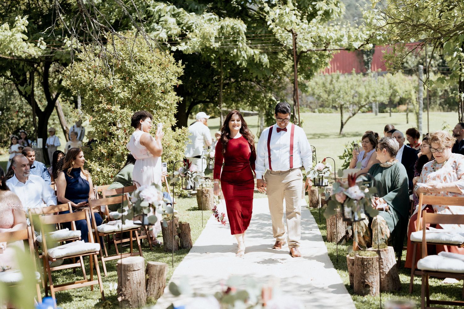 A couple walks down a decorated outdoor aisle at a rustic garden wedding ceremony with guests seated on wooden chairs.