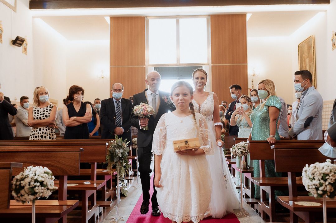 Wedding attendees stand and watch as couple walks down church aisle decorated with white floral arrangements.