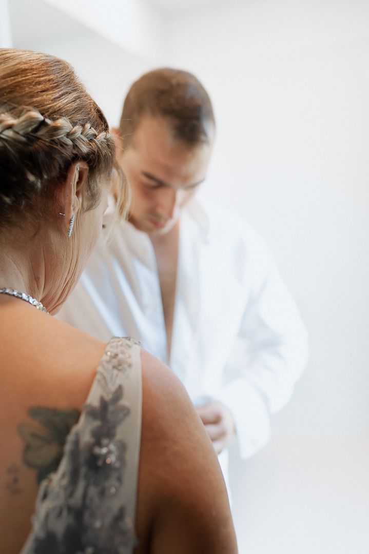A person with braided hair and back tattoo wears silver jewelry while being photographed during a morning wedding preparation.