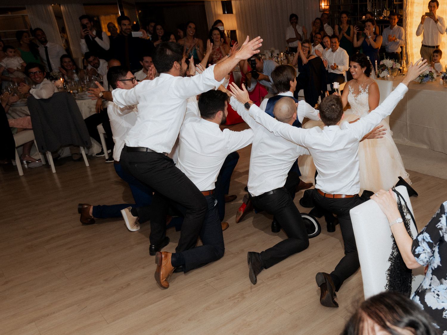 Wedding guests in white shirts perform an energetic dance routine on a wooden floor at a reception celebration.
