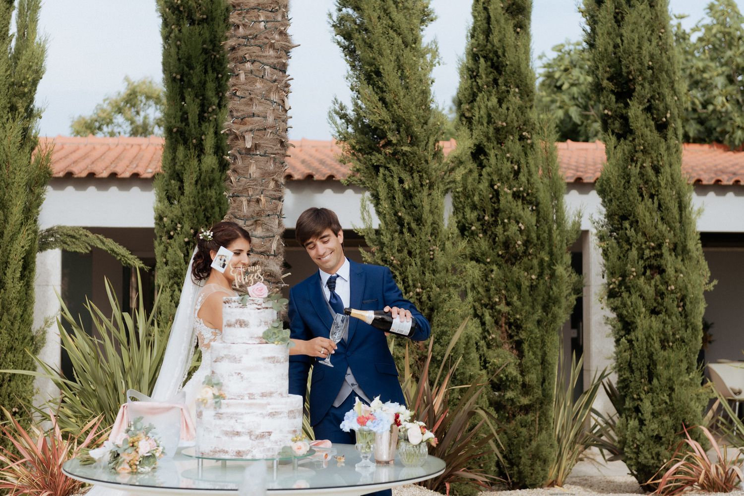 Wedding couple stands at an outdoor table with drinks, surrounded by tall cypress trees and Spanish-style architecture.