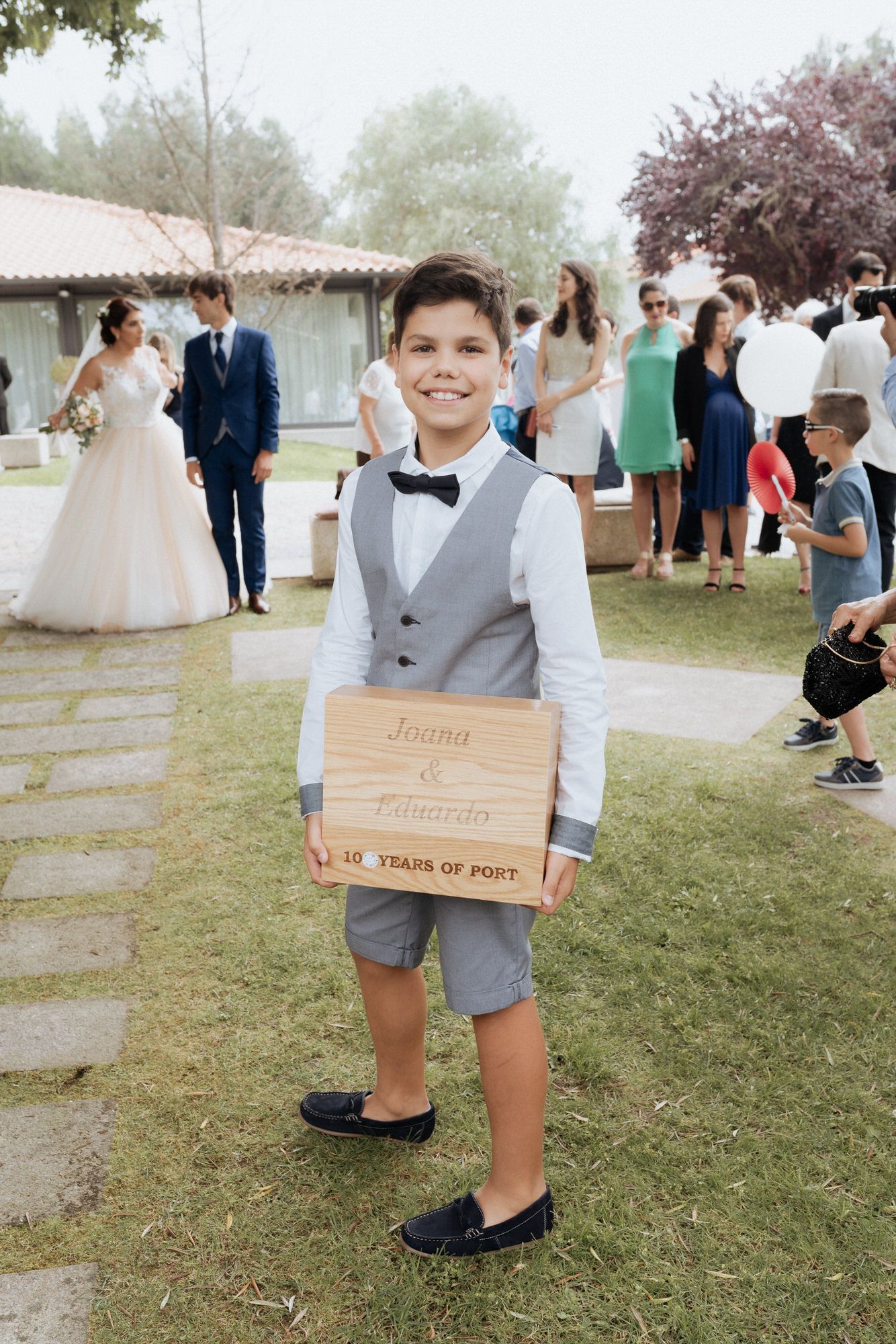 A young attendant in a gray vest and bow tie holds a wooden sign at an outdoor wedding celebration.