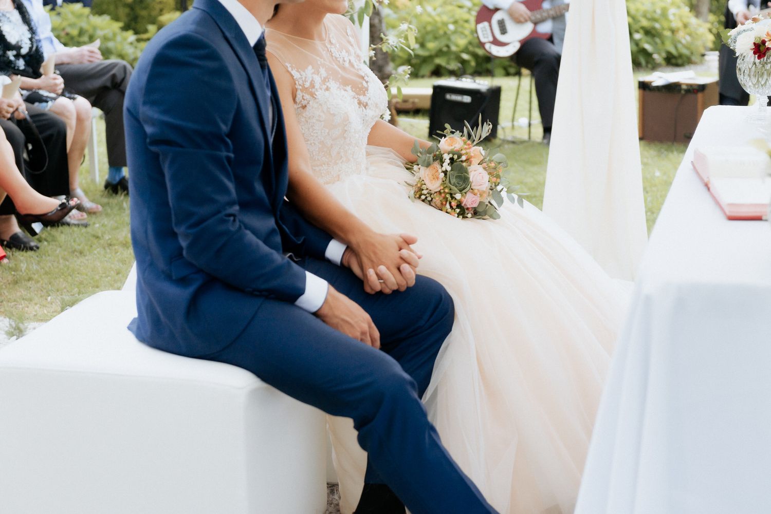 Close-up view of a couple sitting at outdoor wedding ceremony, with groom in navy suit and bride in lace white dress.