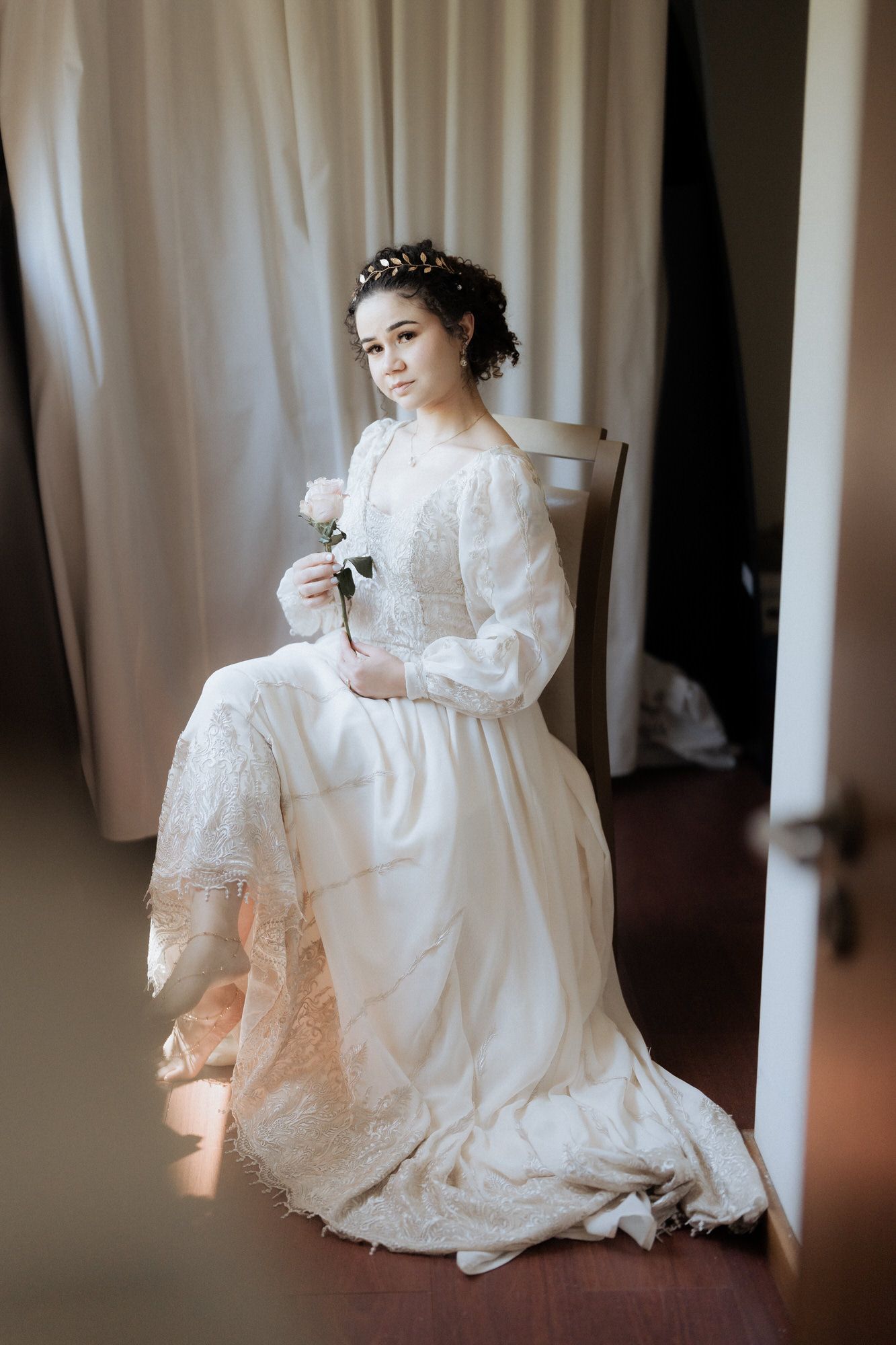 An ethereal bridal portrait of a person in a vintage-inspired white lace gown with puff sleeves holding a single flower.