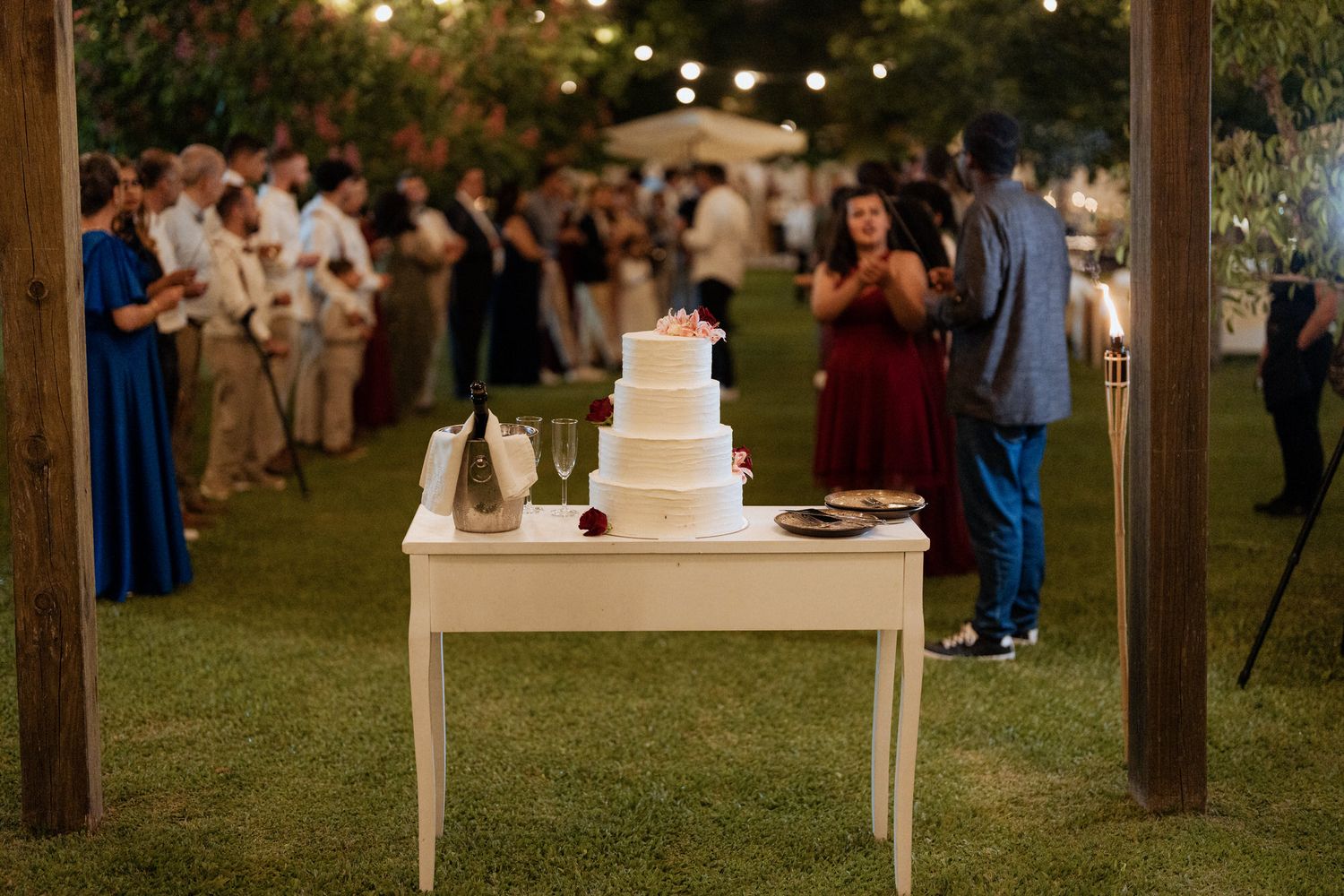 A white wedding cake sits on an elegant table outdoors as guests mingle on the lawn at dusk during a reception.