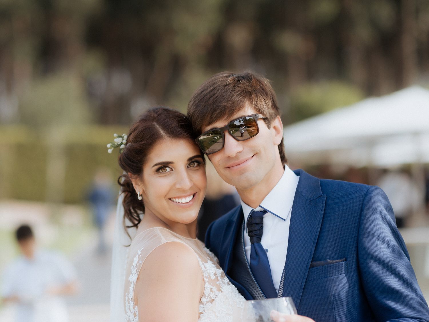 A happy couple in wedding attire smiles together outdoors on a sunny day with trees and white tents in the background.