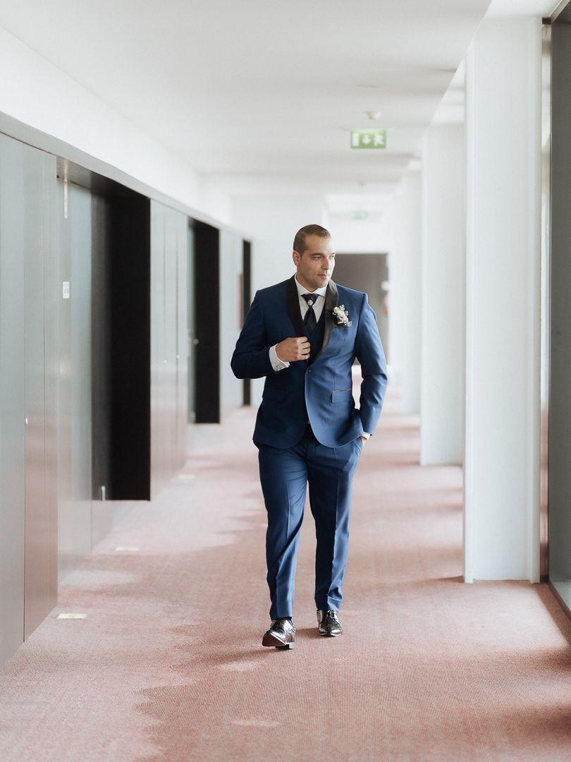 A businessman in a navy blue suit walks confidently down a modern office hallway with carpeted floors.
