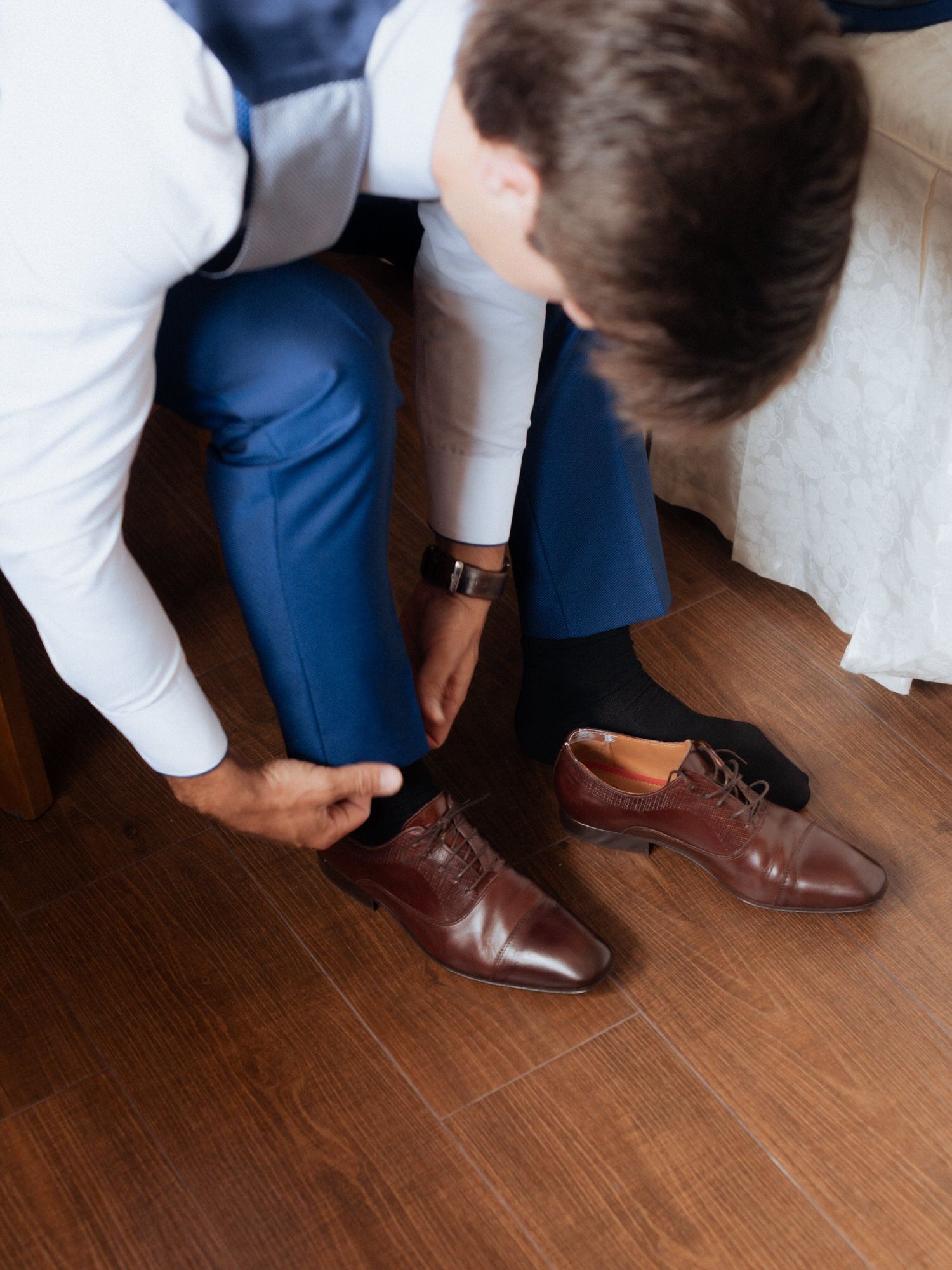 Someone wearing blue pants and a white shirt puts on brown leather dress shoes on a hardwood floor.