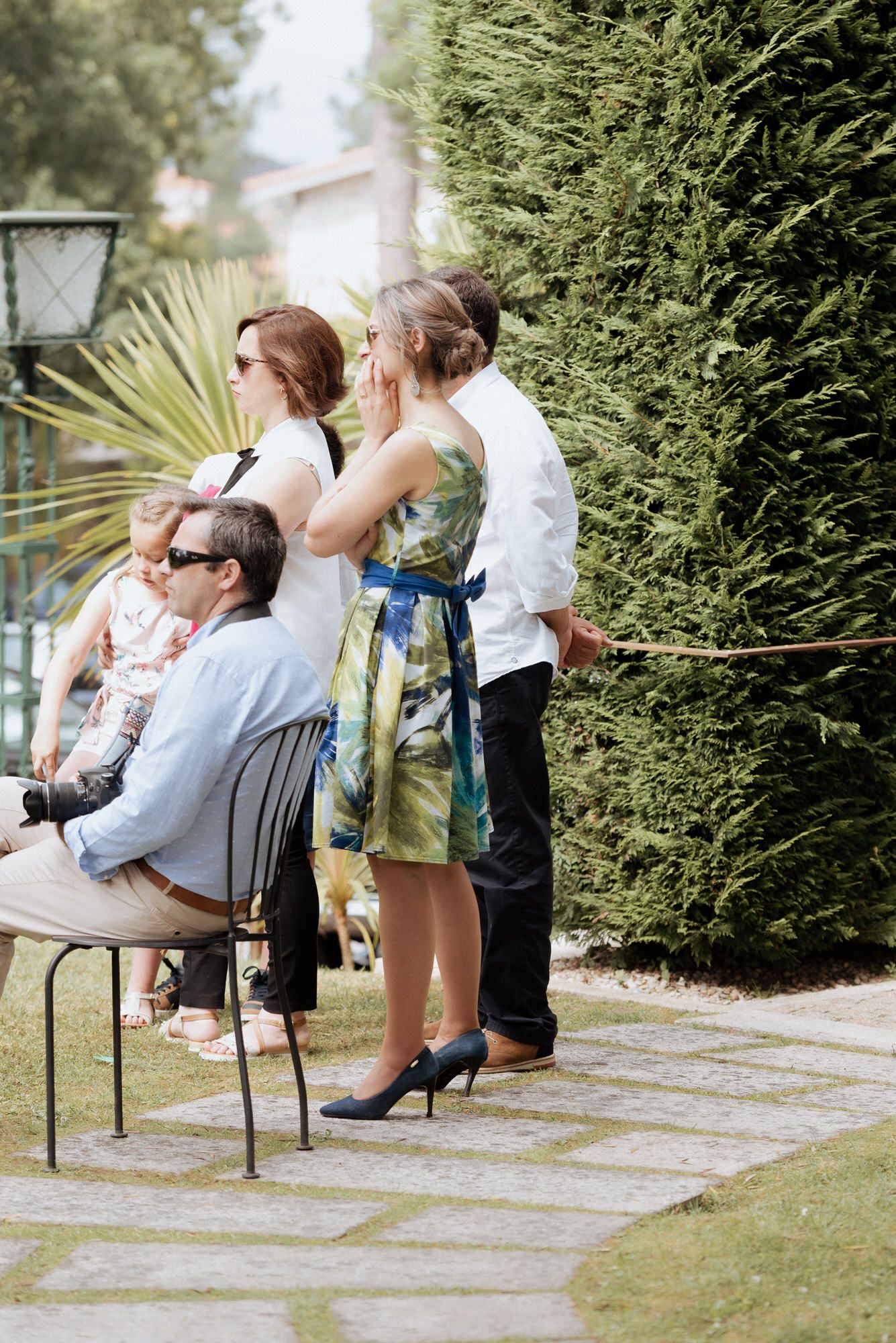 People gathering in a garden setting with lush greenery and paved path on a sunny day.
