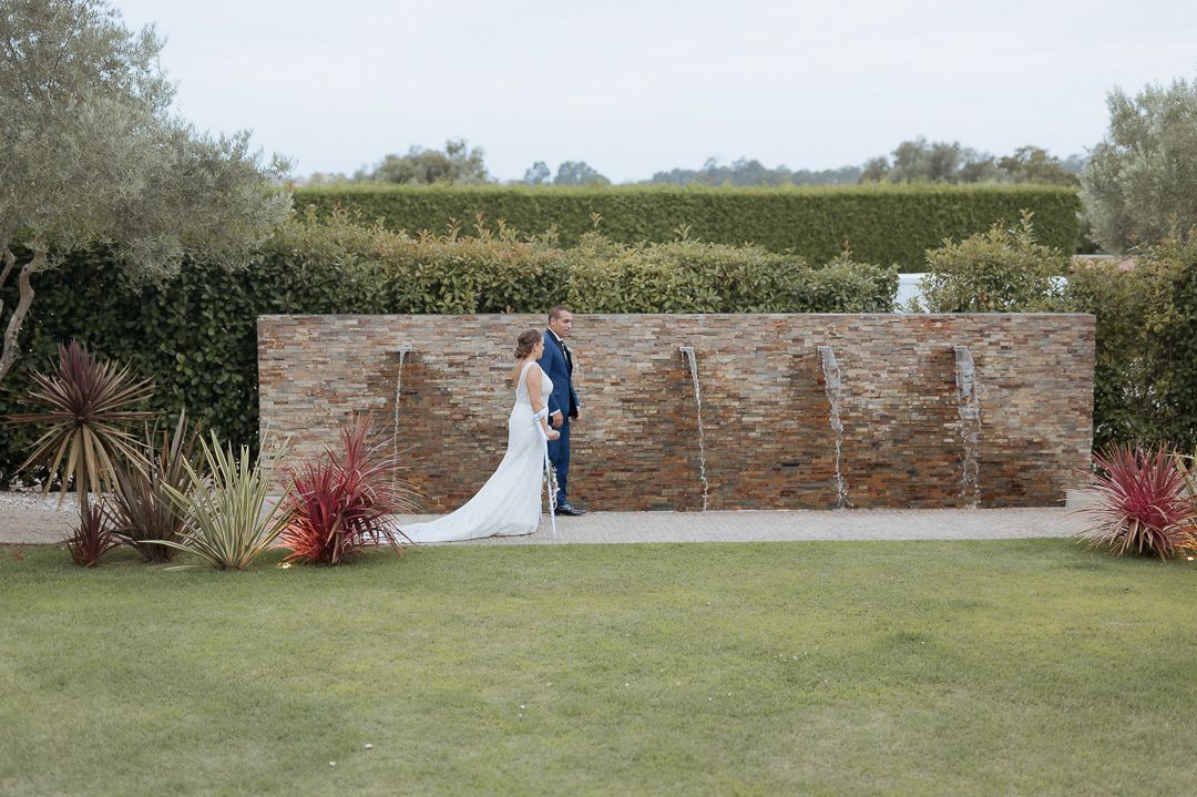 A couple in formal wedding attire stands against a modern stone wall backdrop in an outdoor garden setting.