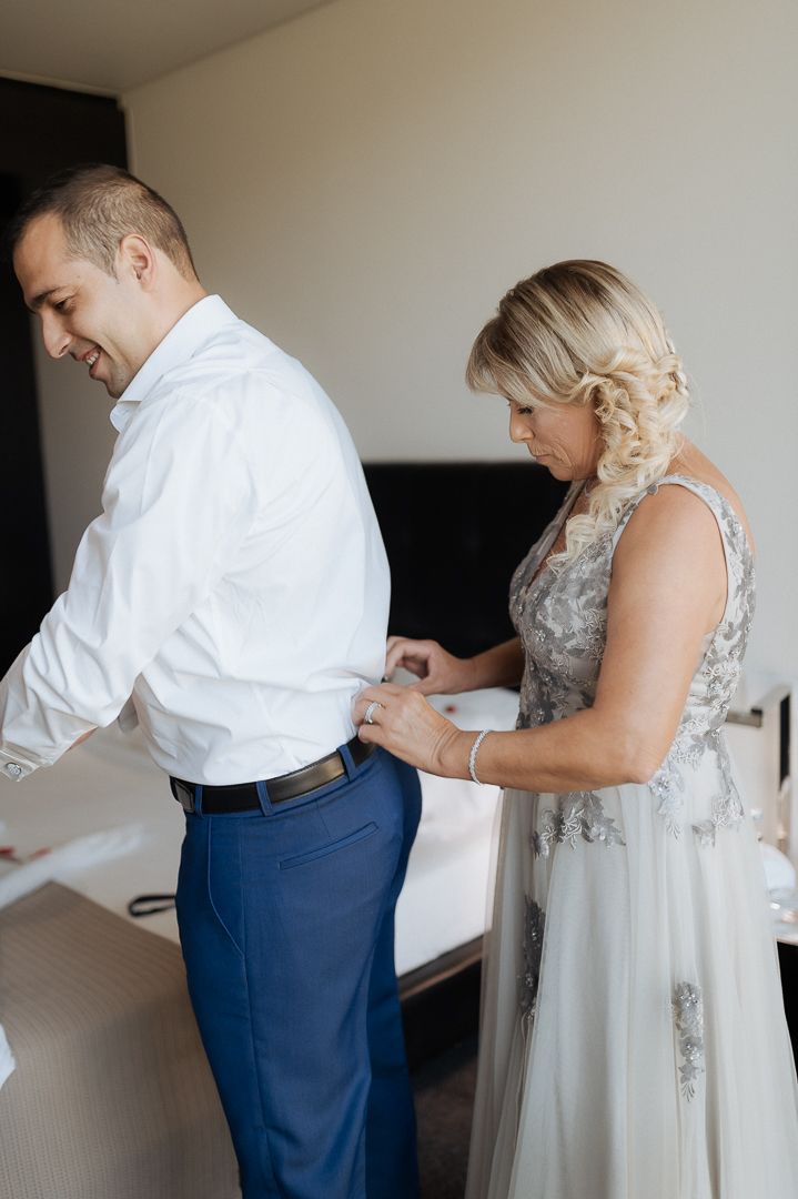 A helpful person assists with adjusting formal attire during wedding preparations in a hotel room.