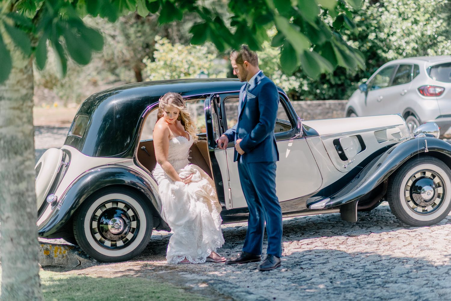 O casal de noivos está ao lado de um carro clássico vintage sob árvores verdes e frondosas.