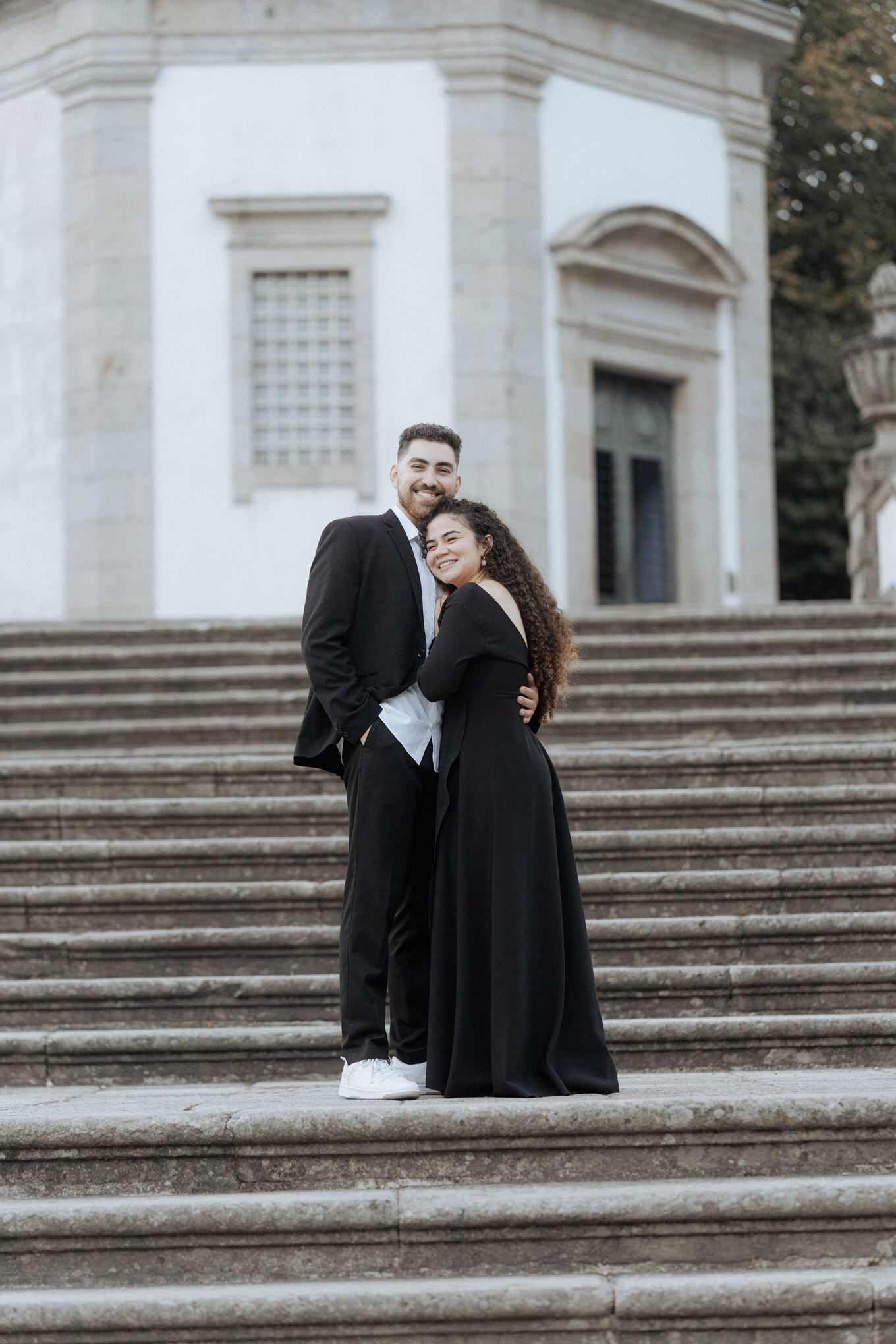 Casal elegante em traje preto formal se abraça nos grandes degraus de pedra branca de um edifício clássico. Namoro no Bom Jesus de braga.