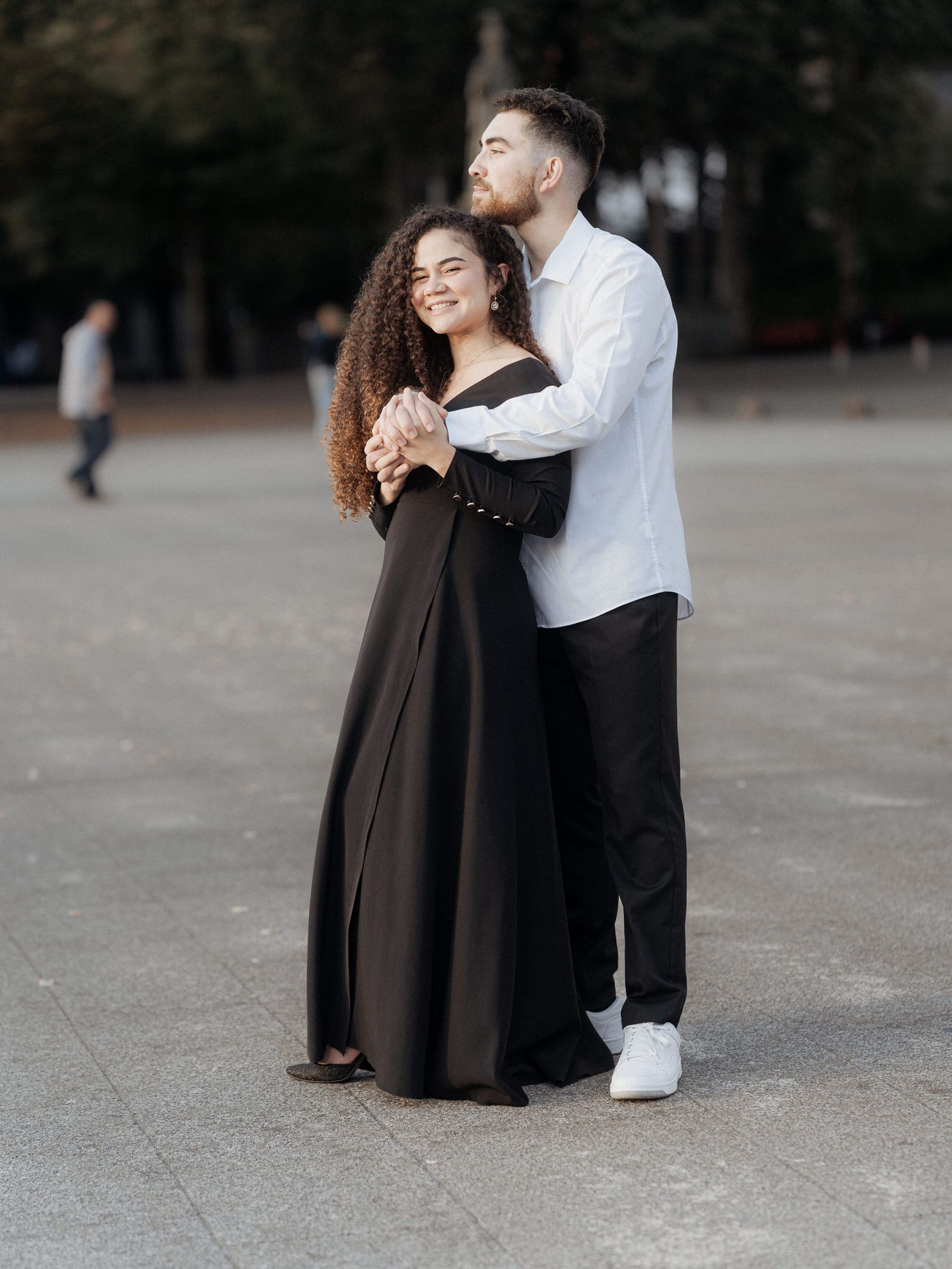 Um casal romântico se abraça ao ar livre, com uma mulher em um longo vestido preto e um homem em uma camisa branca e calças escuras.