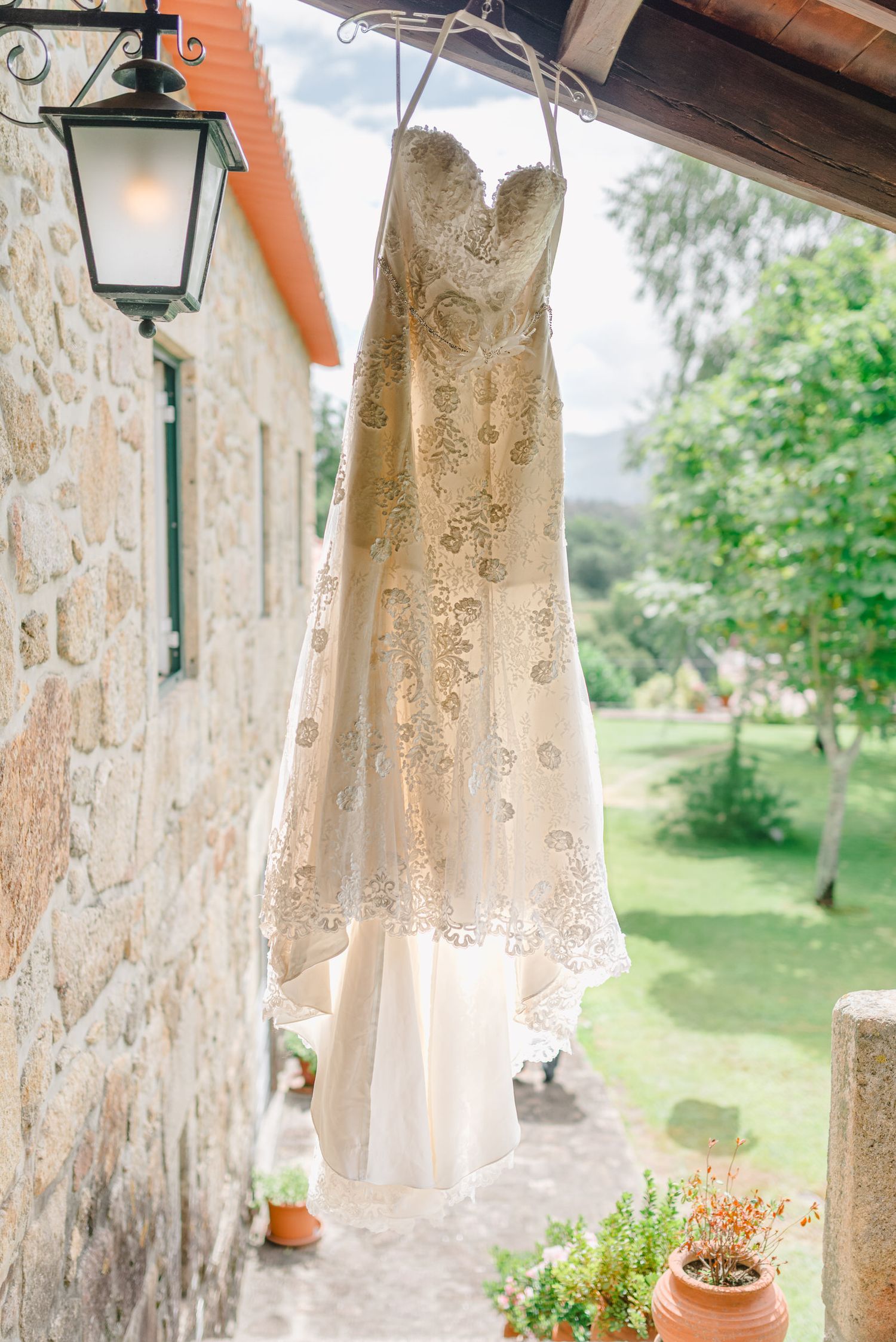 Vestido de noiva ornamentado em renda branca pendurado do lado de fora de uma vila de pedra com vista para o jardim.