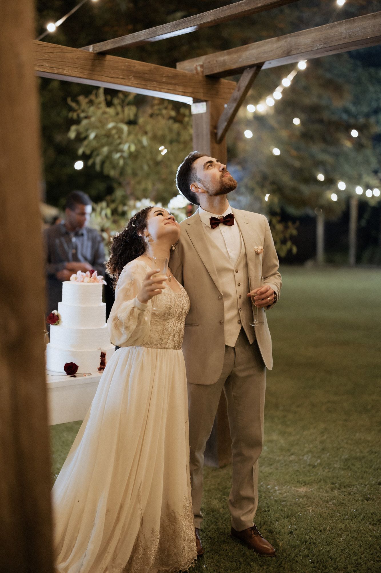 Two people celebrate at an outdoor wedding reception under string lights and wooden beams.