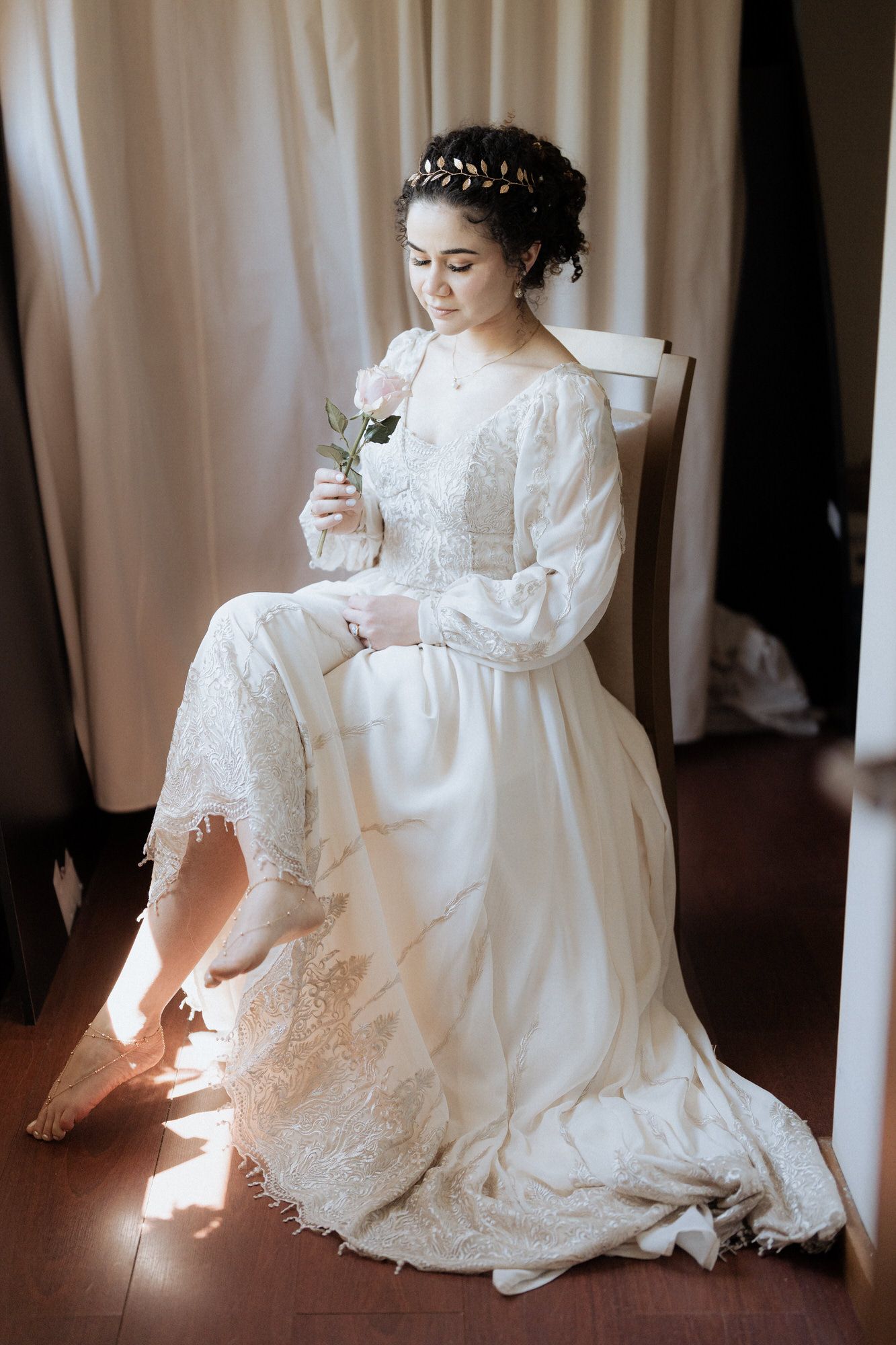 A bride in a vintage-inspired white lace gown with long sleeves sits elegantly holding a flower in soft window light.