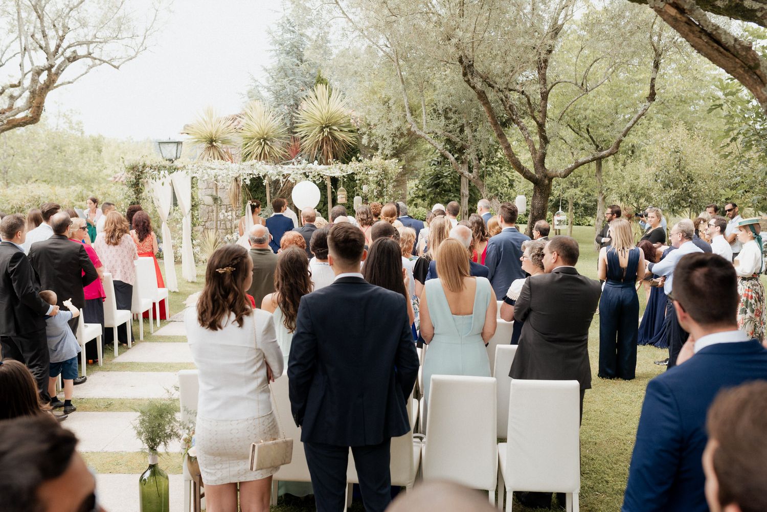 Guests gather outdoors under blooming trees for a spring wedding ceremony in a serene garden setting.