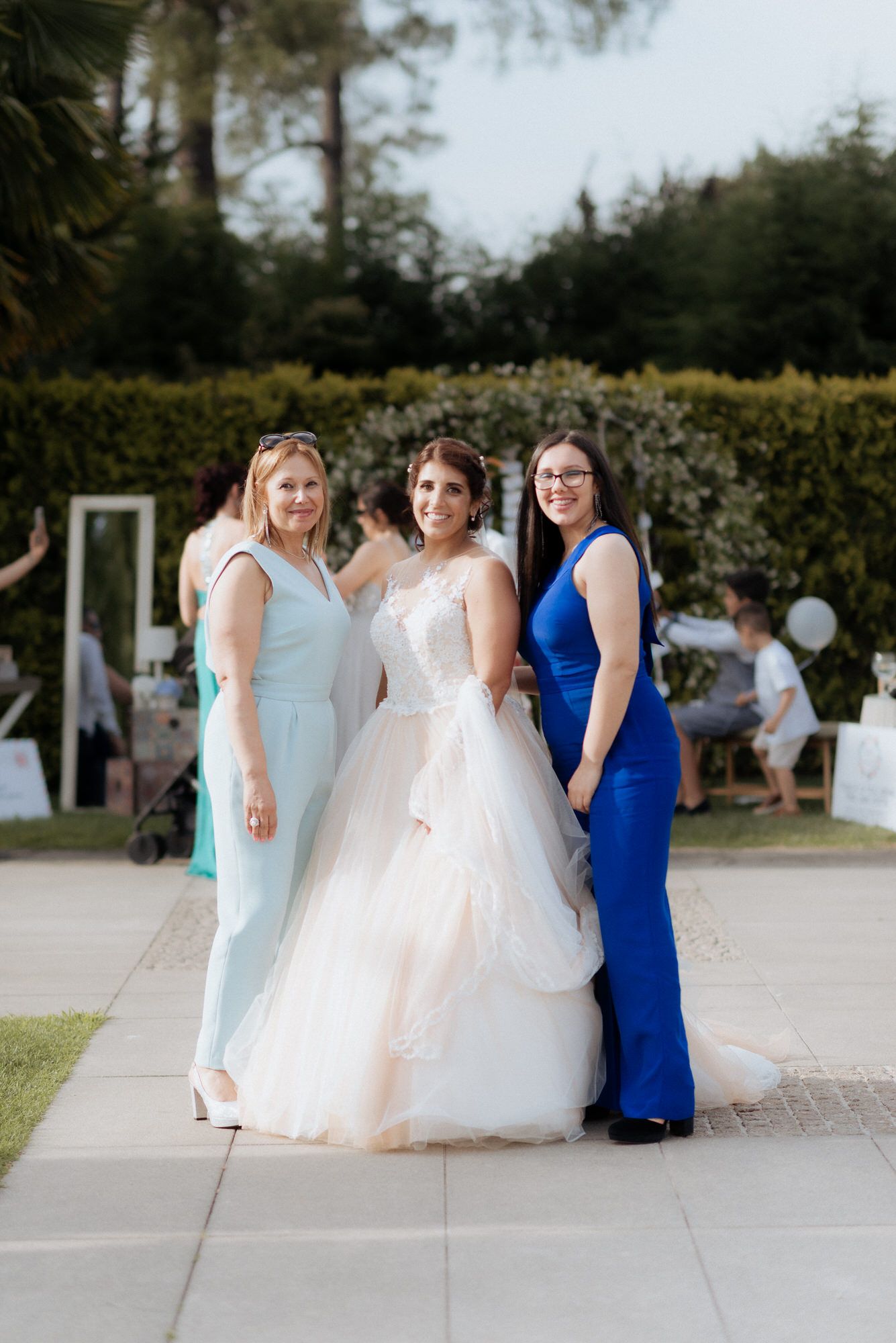 Three women pose at an outdoor wedding venue, with the bride wearing a white ballgown and two guests in blue and mint dresses.