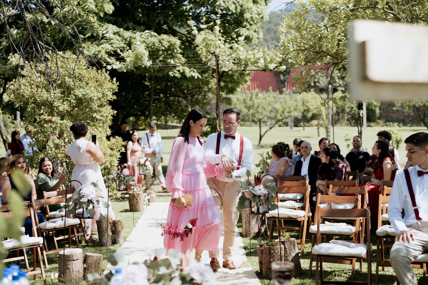 Outdoor garden wedding ceremony with guests seated on white chairs and pink decor along a sunny walkway.