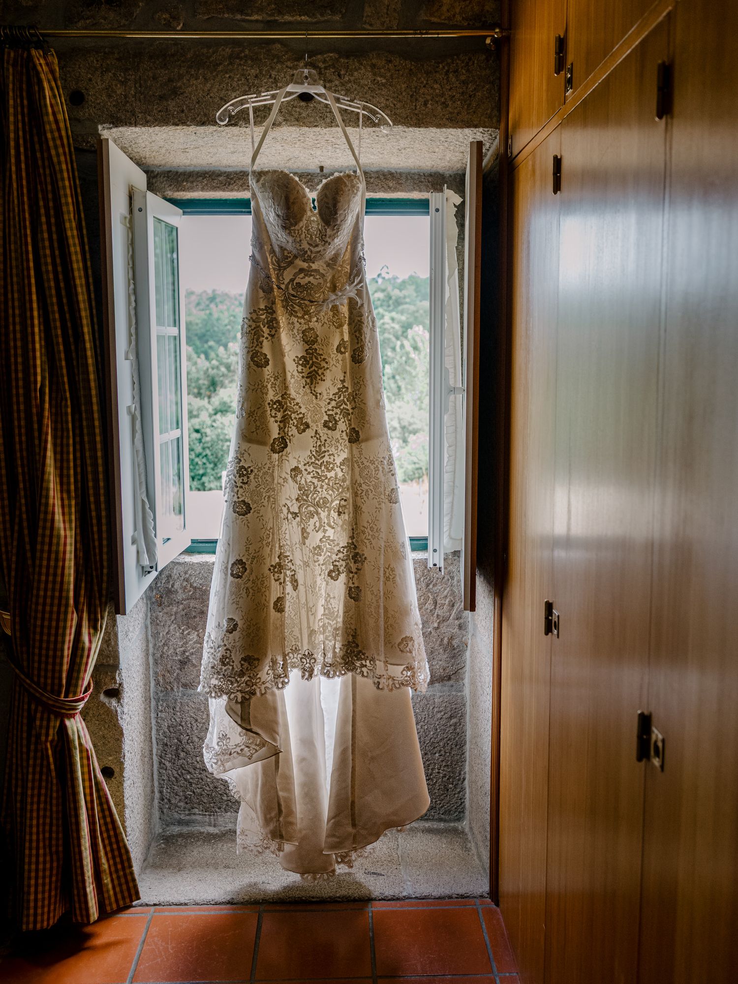 Um elegante vestido de noiva de renda está pendurado na moldura de uma janela, iluminado por luz natural.