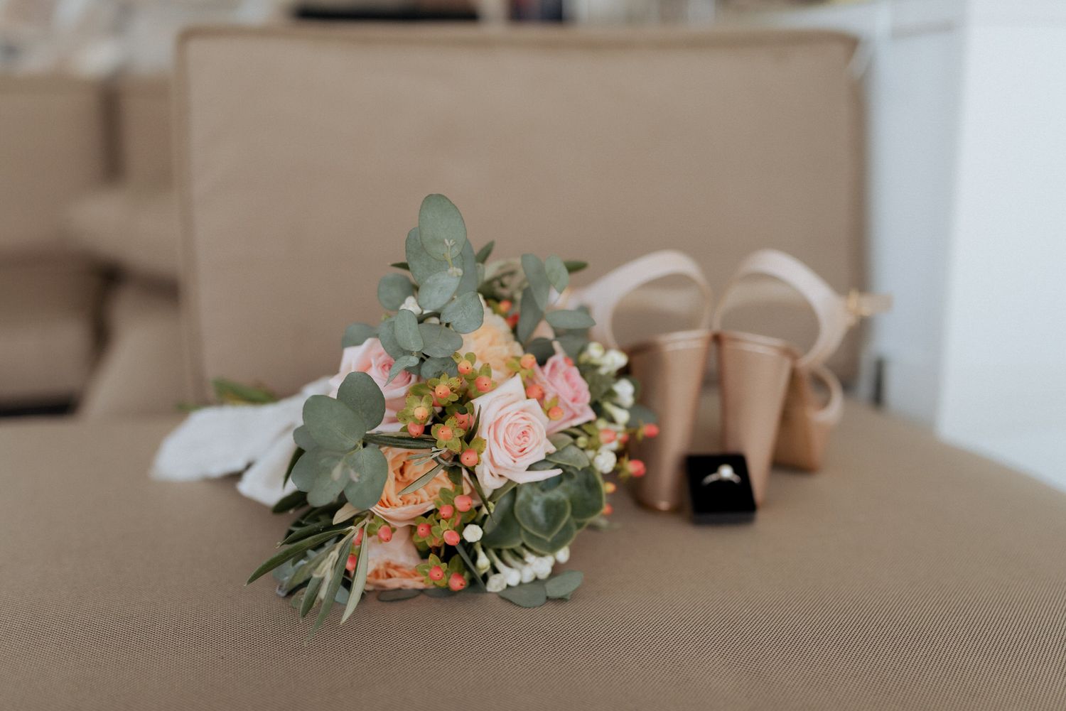 A delicate bouquet of pink roses and eucalyptus leaves sits next to beige wedding shoes on a neutral surface.
