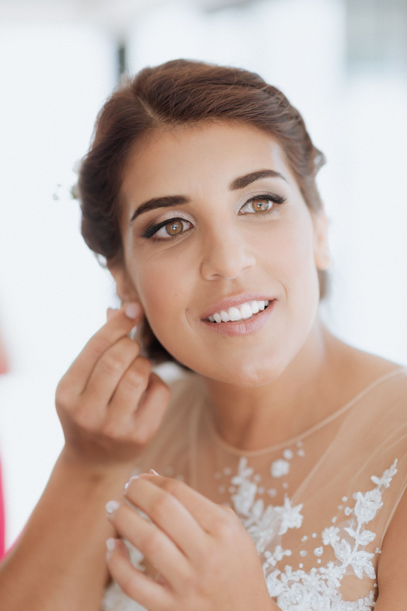A radiant bride in a delicate white lace dress with sheer illusion neckline smiles while getting ready on her wedding day.