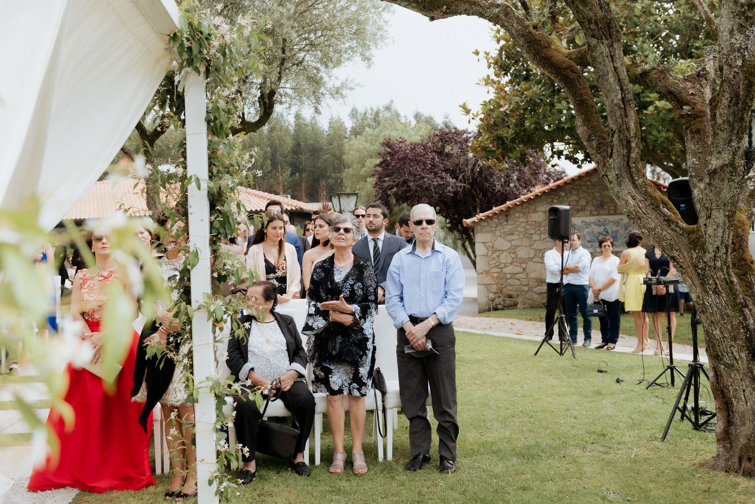 Guests mingling on a sunny lawn at an outdoor garden wedding with trees and stone walls in the background.