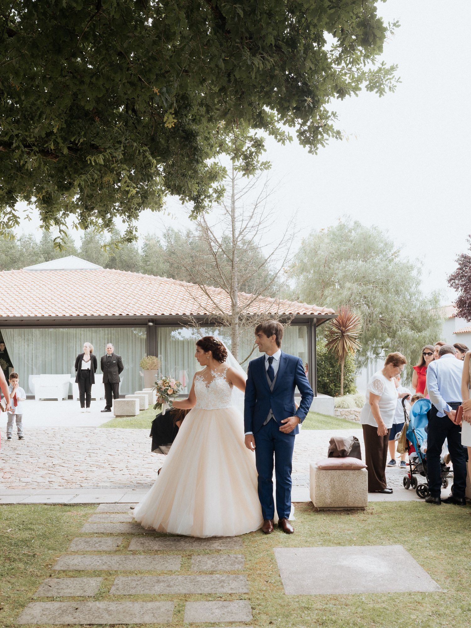 A couple in wedding attire walking together on a stone path outside a Spanish-style venue with guests in the background.