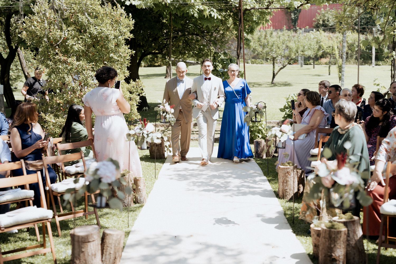 Wedding guests gather outdoors on wooden benches beneath trees during a sunny garden ceremony.