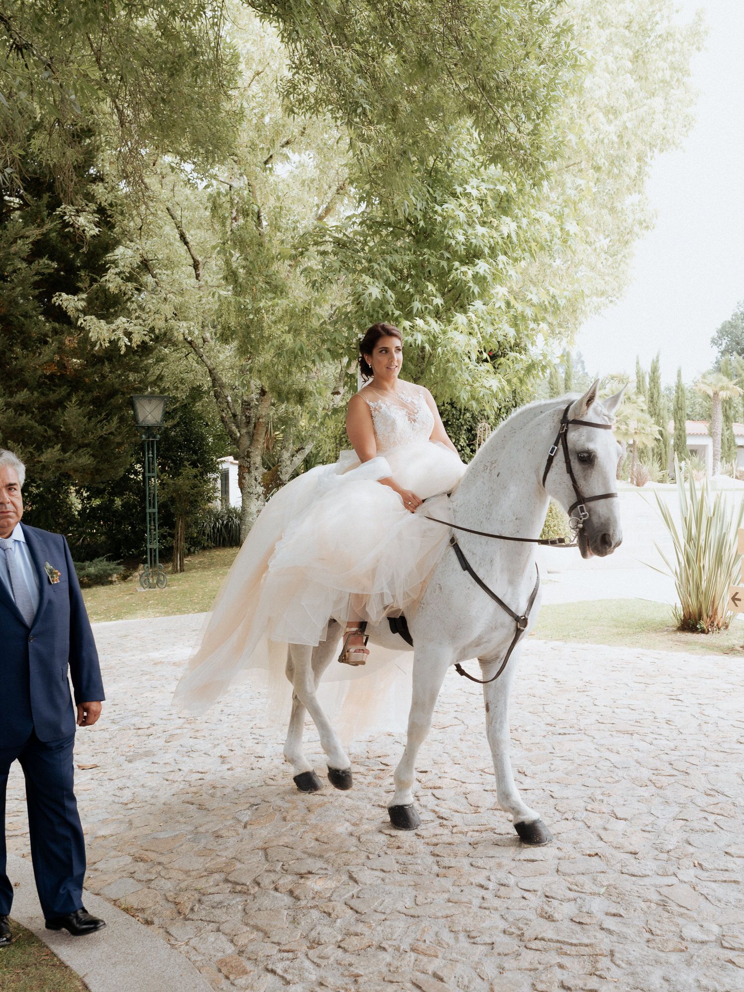 A person in a flowing white wedding dress sits elegantly atop a majestic white horse on a stone path while someone watches nearby.