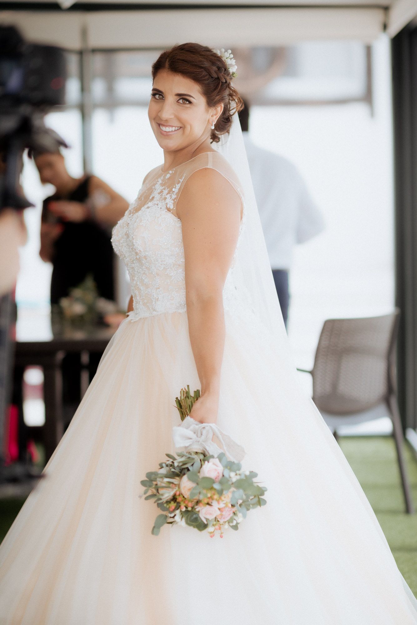 A bride in a white lace and tulle ball gown holds a soft pastel bouquet while smiling at her wedding celebration.