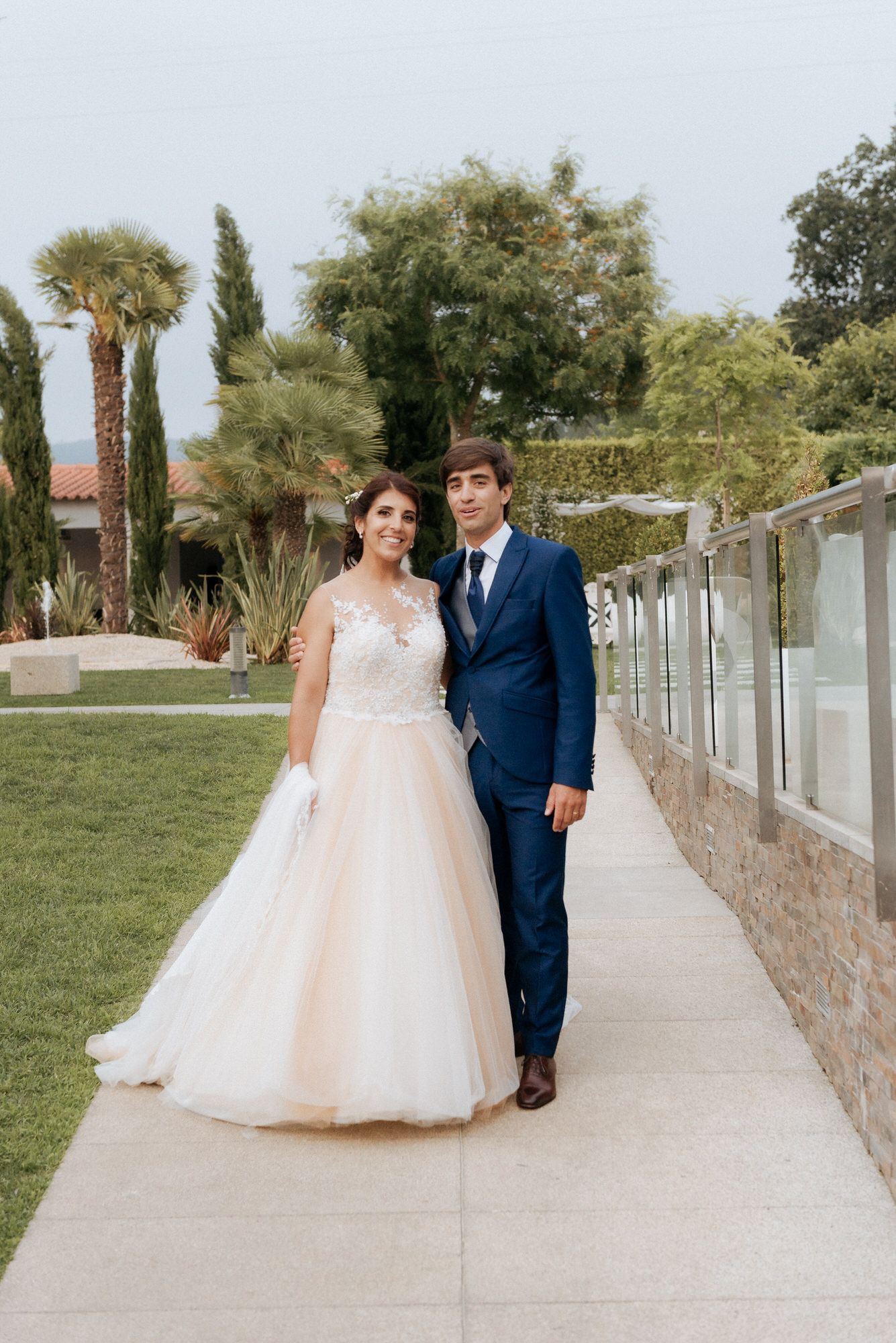 A couple in wedding attire stands together on a concrete pathway bordered by palm trees and landscaping.